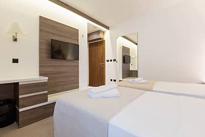 The image shows a modern and minimalist hotel room with two beds, crisp white bedding, wooden accents, a wall-mounted TV, and soft lighting.
