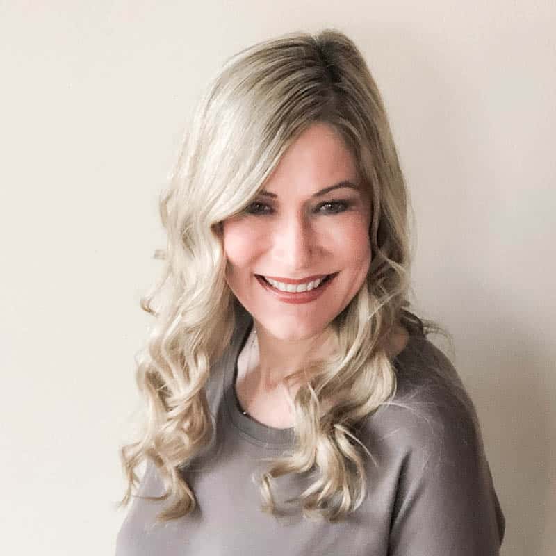 The image shows a smiling person with blonde, curly hair, wearing a grey top, against a plain, light-colored background.