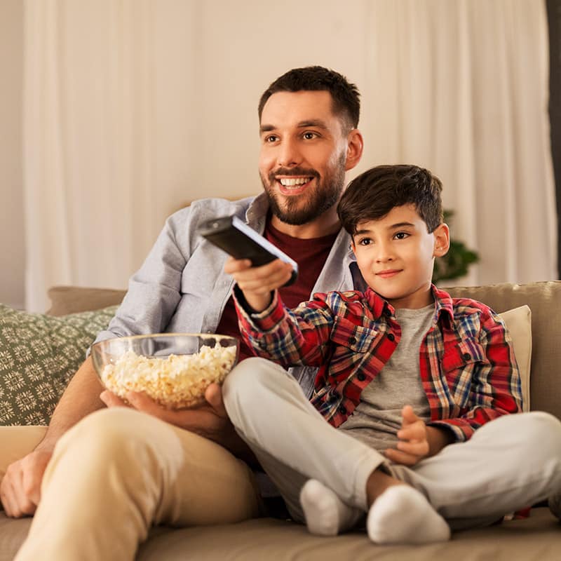 A person and a child are sitting on a sofa, holding a remote control with a bowl of popcorn, smiling and watching TV together.