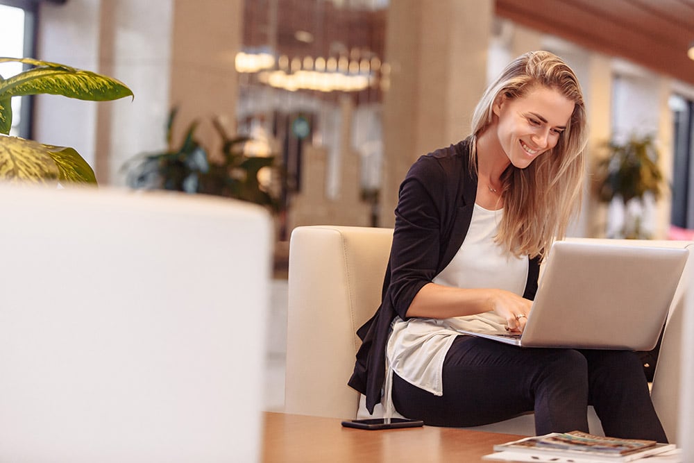 A smiling person is working on a laptop in a comfortable lobby with warm lighting, plants nearby, and a phone on the table.