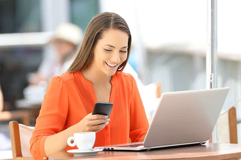 A person sits at a cafe with a laptop, smiling at a smartphone in hand. A coffee cup is on the table. Bright, casual, contemporary setting.