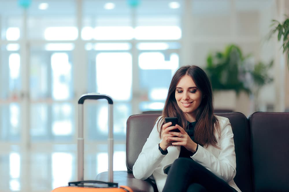 A person sits in an airport lounge, engrossed in their smartphone, with a suitcase beside them, hinting at forthcoming travel or a recent arrival.