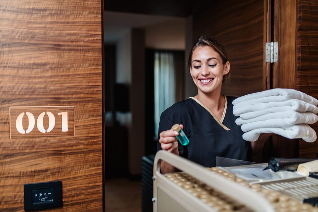 A smiling housekeeper enters a hotel room carrying 3 fresh towels and a small bottle of mouthwash.