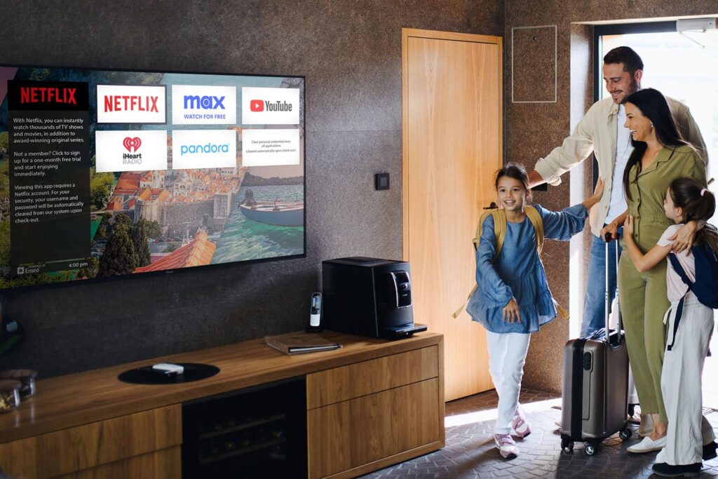 A family of four enters a room with a large TV displaying streaming services. They have luggage, indicating they are starting or ending a trip.